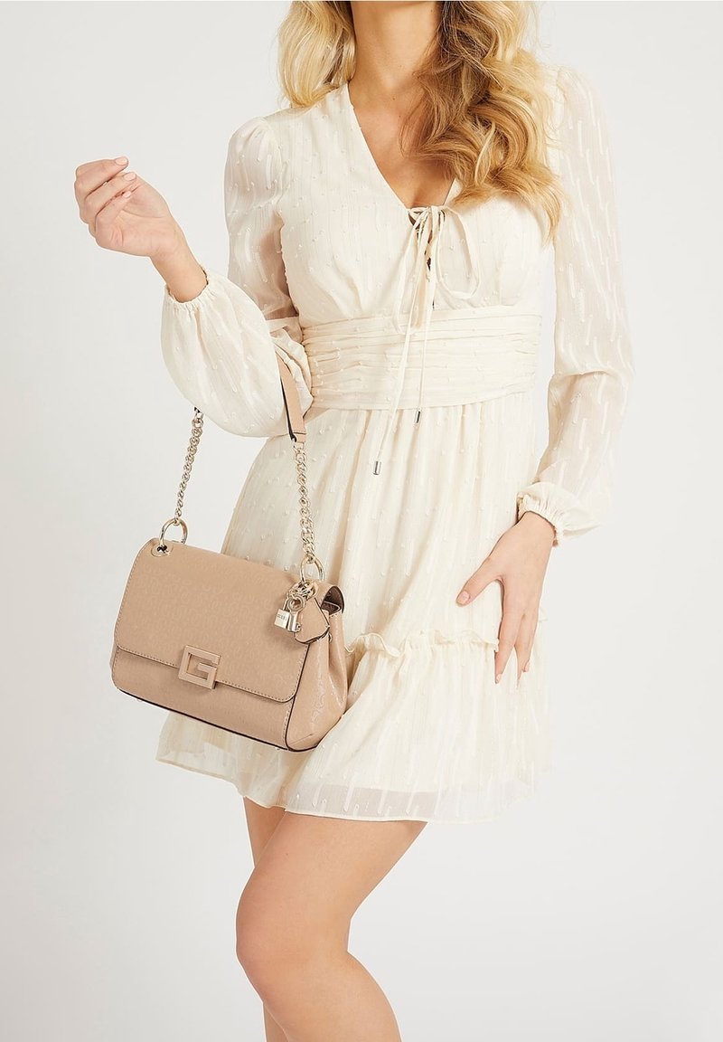 Femme portant une robe crème à manches longues texturée, tenant un sac à main beige avec une bandoulière chaîne dorée et des breloques décoratives.