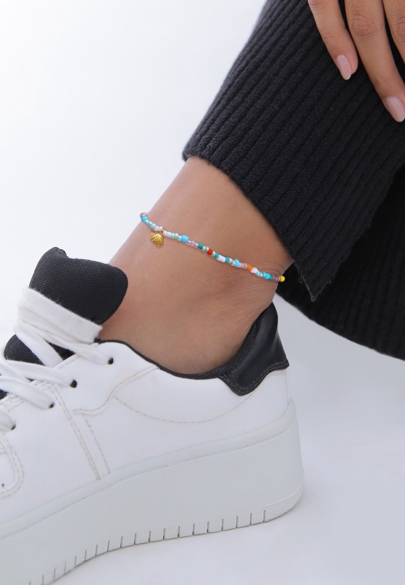 Cheville ornée d'une chevillette colorée en perles avec un petit pendentif en coquillage doré, portant un pantalon côtelé noir et des baskets blanches avec des accents noirs.