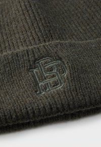 Strickmütze in Olivgrün mit Rippstruktur. Besitzt ein gesticktes Logo in passender Farbe nahe dem Saum. Das Material wirkt weich und warm.