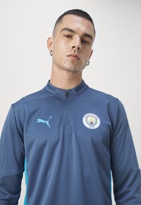 Trenirka Manchester City z zadrgo v temno modri barvi s svetlo modrimi akcenti. Opremljena z logotipom in teksturirano detajlo na rokavih. Narejena iz zračnega materiala.