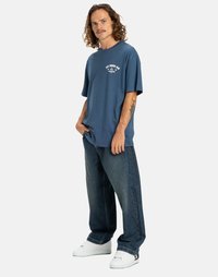 Man med lockigt hår och mustasch iförd en blå DC Shoe Co T-shirt, lösa mörka jeans, vita sneakers, står med händerna i fickorna.