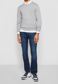 Uomo con un maglione grigio chiaro sopra una camicia bianca, jeans blu e sneaker bianche e nere, in piedi davanti a uno sfondo grigio chiaro.