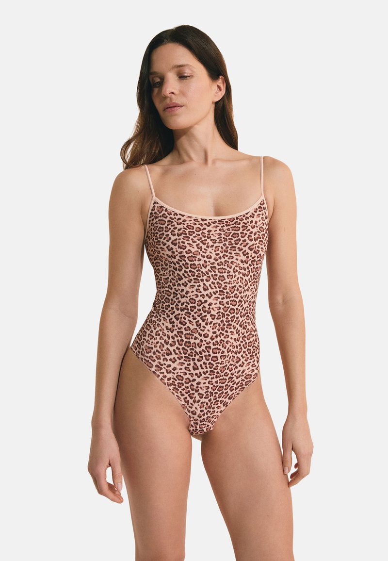 Vrouw met lang bruin haar, gekleed in een mouwloos bodysuit met luipaardprint en dunne schouderbandjes, staande tegen een effen achtergrond.