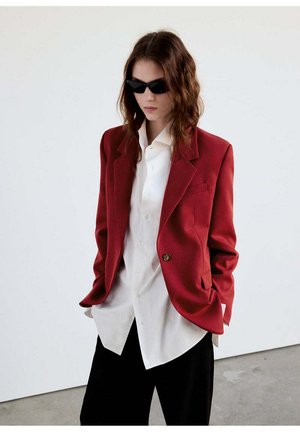 Joven mujer con gafas de sol negras, blazer rojo, camisa blanca abotonada y pantalones negros, de pie con las manos en los bolsillos contra una pared lisa.