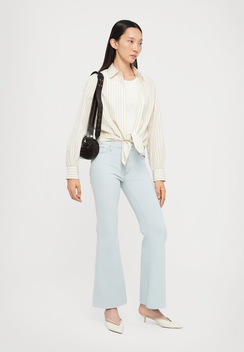 Hellblaue ausgestellte Jeans, beige und weiß gestreiftes Hemd mit Knöpfen, am Bauch gebunden, elfenbeinfarbenes Oberteil darunter, kleine schwarze runde Umhängetasche, neutrale Schuhe.