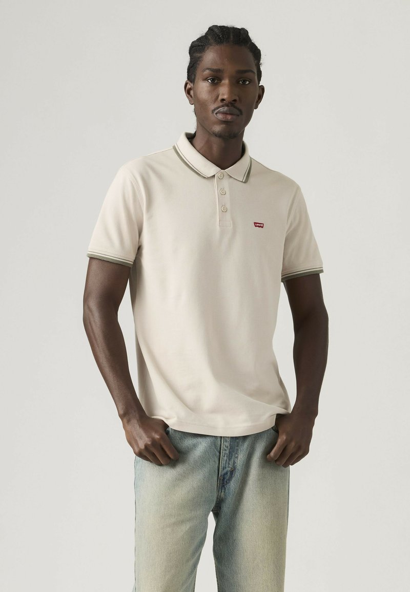 Homme aux cheveux tressés portant un polo beige Levi's et un jean bleu clair, debout devant un fond uni clair.