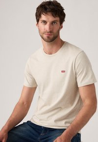 Mann mit kurzbraunem Haar und Bart, der ein beiges Levi's T-Shirt und eine blaue Jeans trägt, sitzt vor einem schlichten hellen Hintergrund.