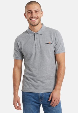 Homme souriant portant un polo gris ellesse et un jean bleu, debout devant un fond blanc uni.
