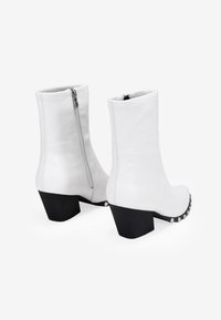 Bottes blanches en cuir mi-mollet avec talons carrés noirs, fermetures éclair latérales et clous décoratifs argentés à l'avant. Design élégant avec une texture lisse.