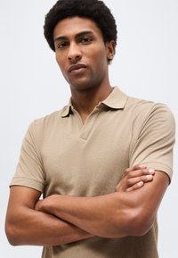 Νεαρός με κοντά σγουρά μαλλιά, φορώντας μπεζ πόλο πουκάμισο, με σταυρωμένα χέρια, κοιτάζοντας με αυτοπεποίθηση μπροστά σε απλό φωτεινό φόντο.