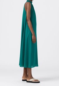 Langes türkisfarbenes Kleid aus fließendem, leichtem Stoff mit hohem Ausschnitt; getragen mit flachen, zweifarbigen Sandalen in minimalistischem Design.