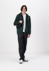 Zöld zipzáras kapucnis pulóver, fehér T-shirt-ra húzva, sötét farmernadrággal és szürke sportcipővel párosítva, egy fekete válltáskával a kezében.