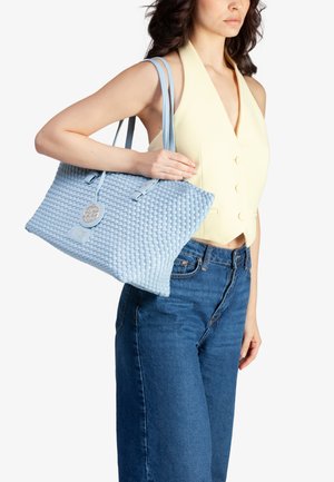 Lichtblauwe geweven tote bag met twee handgrepen, met een cirkelvormig logo en gladde leren accenten; gecombineerd met een gele top en denim spijkerbroek.