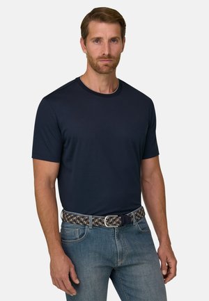 Mężczyzna z krótkimi brązowymi włosami i brodą, ubrany w ciemnoniebieski t-shirt z krótkim rękawem, dżinsy oraz pleciony pasek na jednolitym tle.