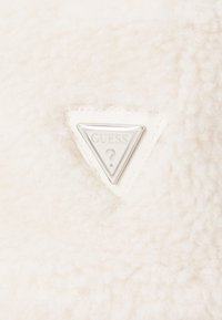 Fehér plüss anyag, textúrált felülettel, középen háromszögletű ezüst "GUESS" logóval kiemelve.