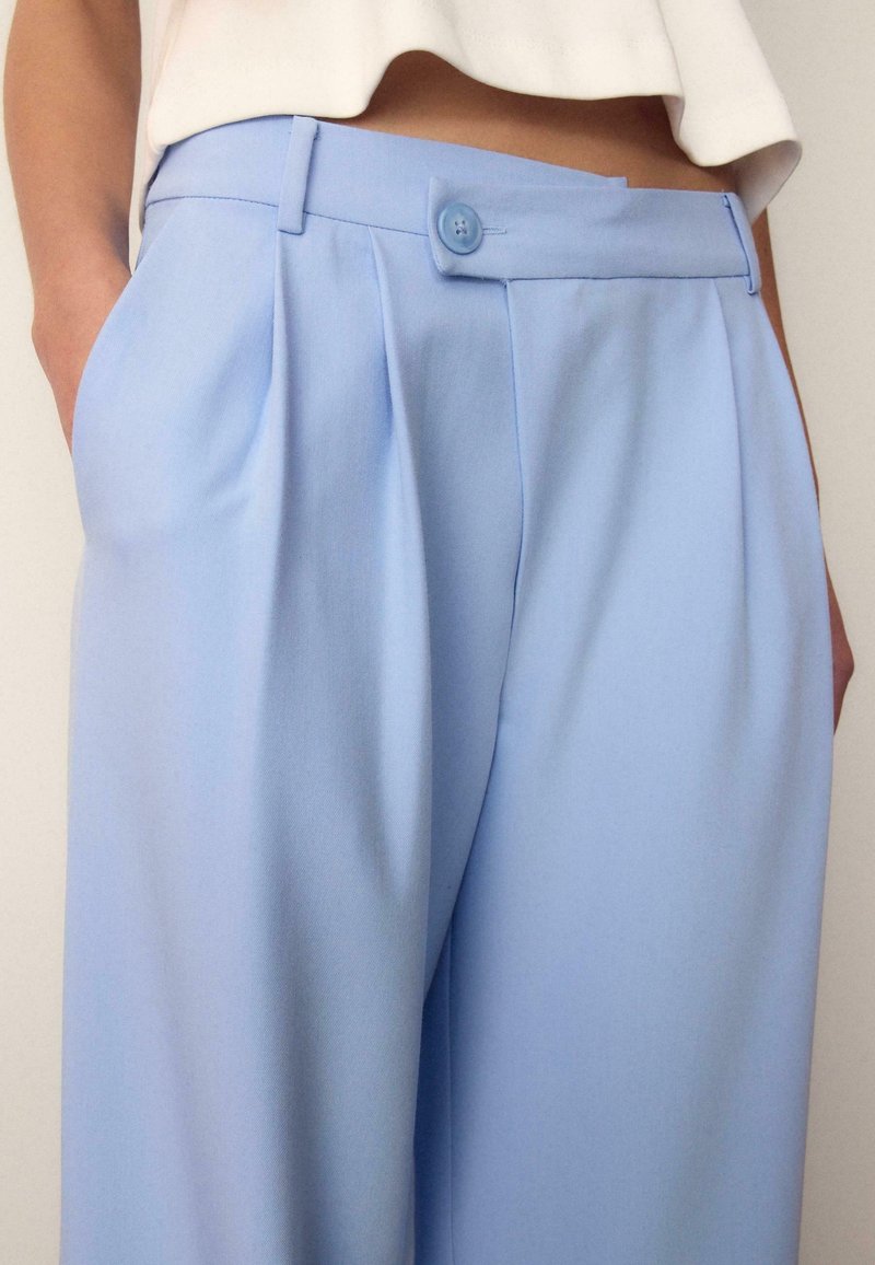 Personne portant un pantalon bleu clair taille haute avec des plis et une fermeture à boutons, mains dans les poches, associé à un top blanc court.