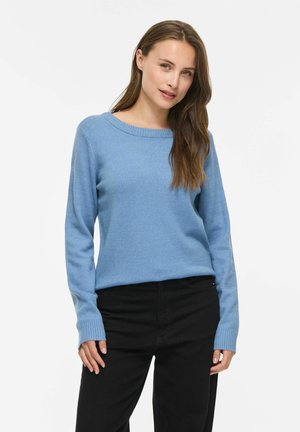 Femme aux longs cheveux bruns portant un pull bleu clair à manches longues et un pantalon noir, debout devant un fond blanc uni.