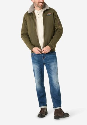 Olivgrüne Bomberjacke mit einem Kunstfellkragen, Reißverschluss vorne und Patch-Details. Kombiniert mit blauen Jeans und braunen Schnürstiefeln.