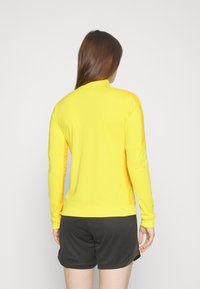 Long-sleeve yellow sports top with a high collar, smooth texture, and fitted design, paired with black shorts featuring a subtle pattern.