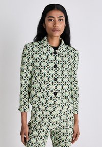 Gemusterte Jacke mit schwarzem Hintergrund, die ein geometrisches Design in Minzgrün und Creme zeigt. Sechs schwarze Knöpfe und ein Kragen.