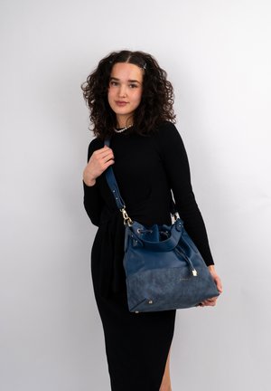 Borsa a secchiello in pelle e suede blu navy con chiusura a coulisse, dettagli in hardware dorato e tracolla removibile, tenuta da una persona in abito nero.