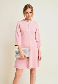 Rosa klänning med hög hals, trekvartslånga ärmar och en dekorativ silvertrim ner längs framsidan. Modellen håller en silverfärgad kuvertväska.