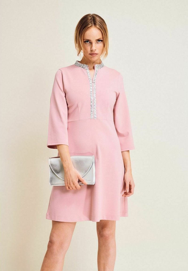 Rosa klänning med hög hals, trekvartslånga ärmar och en dekorativ silvertrim ner längs framsidan. Modellen håller en silverfärgad kuvertväska.