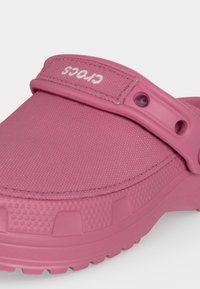 Sabot Crocs rose avec une bride texturée portant le logo blanc "Crocs" et des trous perforés sur un fond clair.