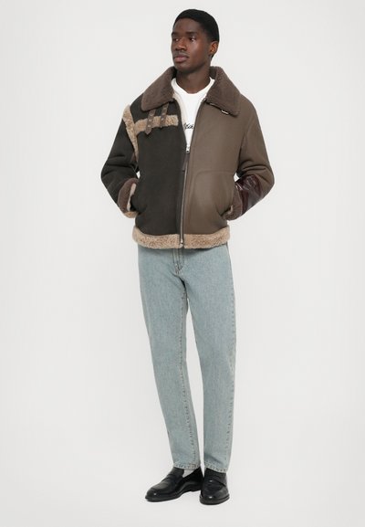 Brauner und beiger Plüschjacke mit großem Kragen, Reißverschluss und kontrastierenden Lederärmeln; kombiniert mit hellblauen Jeans und schwarzen Schuhen.