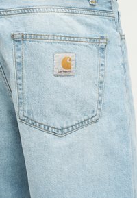 Τσέπη ανοιχτού γαλάζιου τζιν με ορατή ραφή και ραμμένη ετικέτα λογότυπου Carhartt σε κίτρινο και άσπρο.