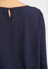 Námořnická modrá blůza s klíčovou dírkou na zadní straně, vyrobená z hladkého materiálu s jemným leskem a minimálními švy.