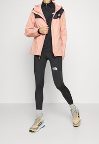 Veste imperméable rose clair avec une section supérieure noire, associée à des leggings noirs et des chaussures de sport beiges avec des accents bleus.
