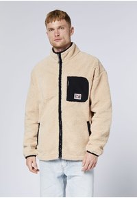 Mann trägt eine beige Fleecejacke mit schwarzer Brusttasche mit Reißverschluss und hellblauen Jeans, steht vor einem schlichten hellen Hintergrund.