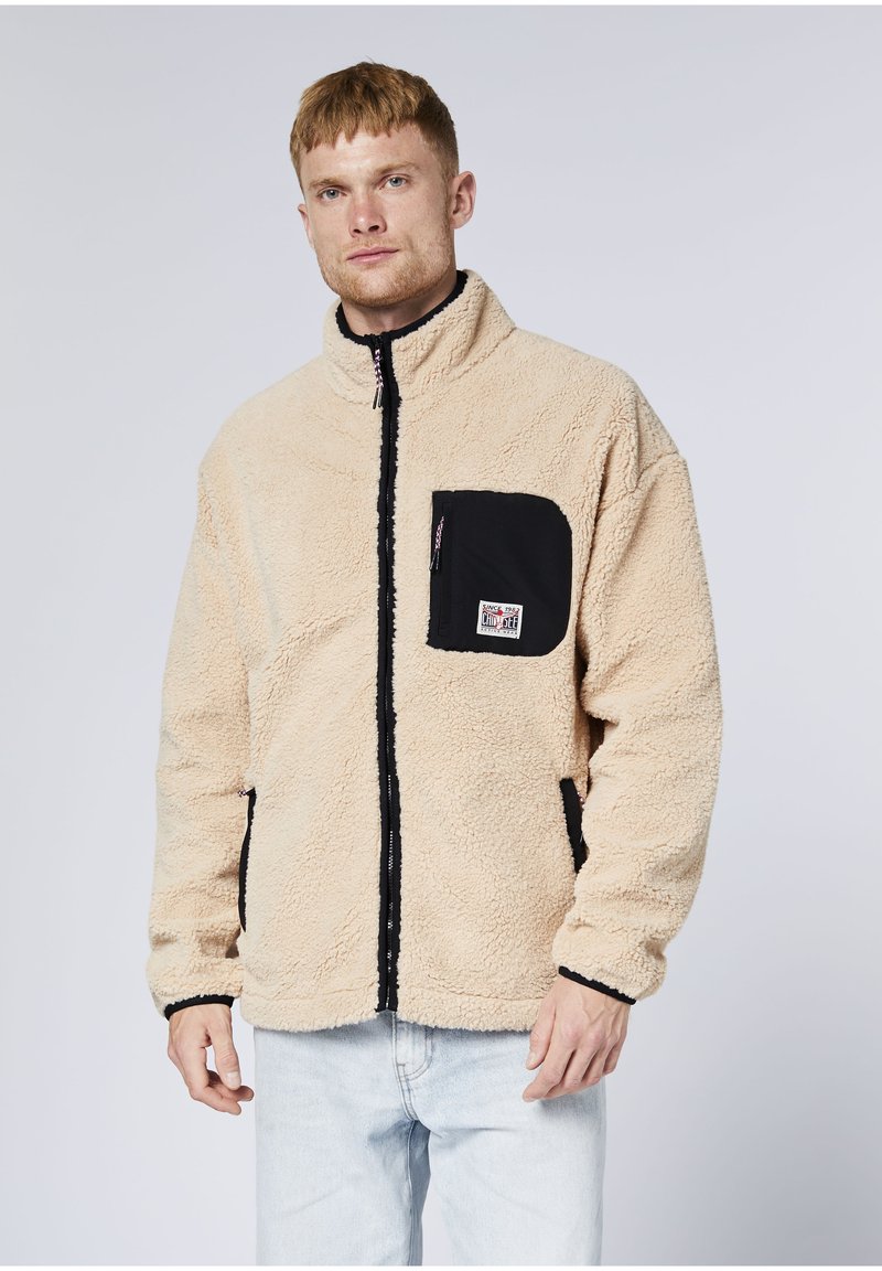 Mann trägt eine beige Fleecejacke mit schwarzer Brusttasche mit Reißverschluss und hellblauen Jeans, steht vor einem schlichten hellen Hintergrund.