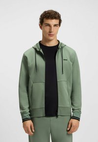 Light green zip-up hoodie with a black logo, front pockets, and drawstring hood. Worn over a black T-shirt, features black accents on cuffs.