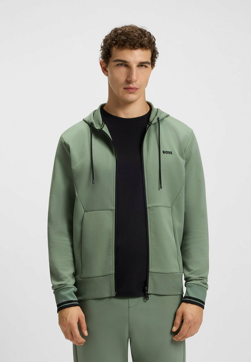 Light green zip-up hoodie with a black logo, front pockets, and drawstring hood. Worn over a black T-shirt, features black accents on cuffs.