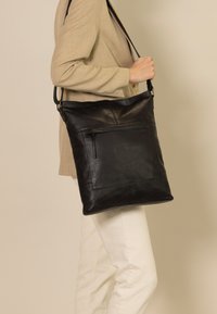 Borsa a tote nera in pelle con una superficie liscia, dotata di una tasca frontale con zip e di una tracolla regolabile. Forma rettangolare semplice.