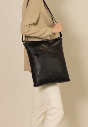 A person wears a light beige blazer and holds a large black leather bag with a front zipper pocket, set against a neutral background.