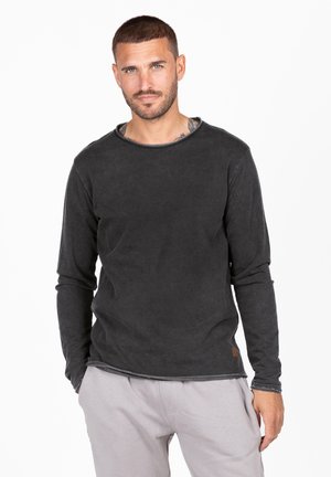Camisa negra de manga larga con cuello redondo, que presenta una apariencia texturada y ligeramente desgastada, y un pequeño acento de cuero en el dobladillo.