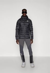 Juoda pūsta striukė su horizontaliu siuvimu ir kapišonu, derinama su pilkomis aptemptomis kelnėmis su atspindinčiomis detalėmis ir juodais intarpais bei baltais sportbačiais.