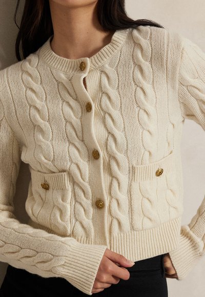 Roomkleurige kabelgebreide cardigan met gouden knopen en twee zakken aan de voorkant, gedragen door een persoon met donker haar en een zwarte broek.