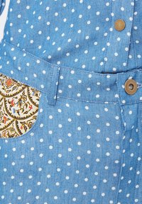 Tela de mezclilla azul con lunares blancos, botones a presión de latón y un bolsillo forrado con tela de paisley amarilla.
