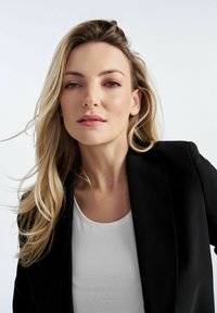 Czarna marynarka na białej bluzce, o dopasowanej sylwetce i gładkim materiale. Długie blond włosy opadają w miękkich falach. Neutralne tło.