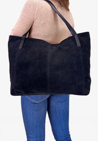 Suuri musta mokkanahasta valmistettu kassi, jossa on nahkaosat ja kaksoisvetoketjut, pehmeällä pinnalla ja tyylikkäällä, minimalistisella muotoilulla.