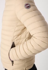 Beige puffer jacket with horizontal quilting, smooth texture, and a side zipper pocket. Features a circular Colmar logo on the sleeve.