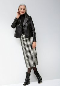 Schwarze Lederbikerjacke über einem eng anliegenden schwarzen Rollkragenpullover, kombiniert mit einem knielangen Hahnentrittrock und schwarzen Stiefeletten.