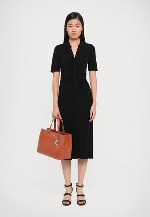 Robe noire à boutons avec des manches courtes et des détails froncés. Modèle tenant un sac à main orange texturé avec deux poignées. Sandales noires à lanières.