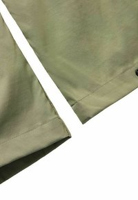 Närbild av olivgrönt tyg med synliga sömmar och en svart knapp på kanten, sannolikt en del av ett plaggets ärm eller byxben.