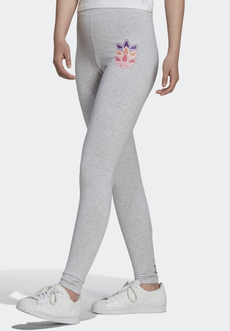 Person som bär grå leggings med en färgglad lövlogotyp på låret och vita sneakers, som står med benen korsade.