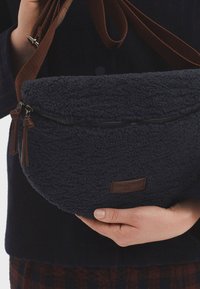 Borsa in pile blu navy con forma arrotondata, dotata di chiusura con zip, dettagli in pelle marrone e una lunga tracolla regolabile.